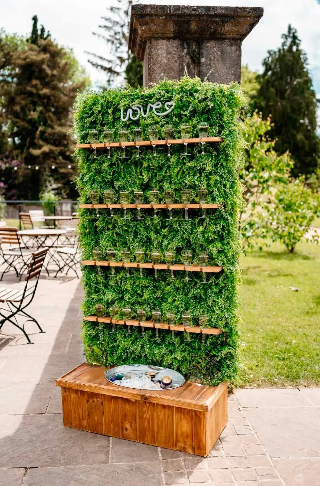 Green foliage champagne wall with wooden shelves of filled flutes in a garden
